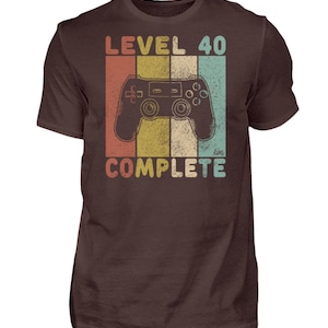 May include: A brown t-shirt with a retro-style graphic of a video game controller and the text "Level 40 Complete".