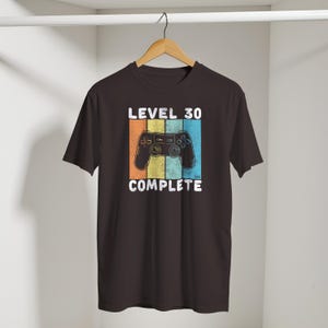 May include: A dark brown t-shirt with a retro design. The design features a video game controller and the words "LEVEL 30 COMPLETE" in white. The background behind the controller is divided into orange, yellow, and blue vertical stripes.
