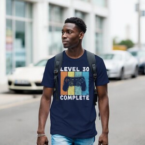 May include: Navy blue t-shirt with a retro video game controller graphic and the text "LEVEL 30 COMPLETE". The design features orange, yellow, and blue stripes. The shirt is made of a soft material.