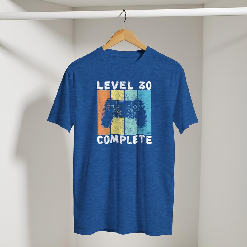 May include: Blue t-shirt with a retro design featuring a video game controller and the text "LEVEL 30 COMPLETE." The design includes orange, yellow, and blue stripes. The shirt is hanging on a wooden hanger.