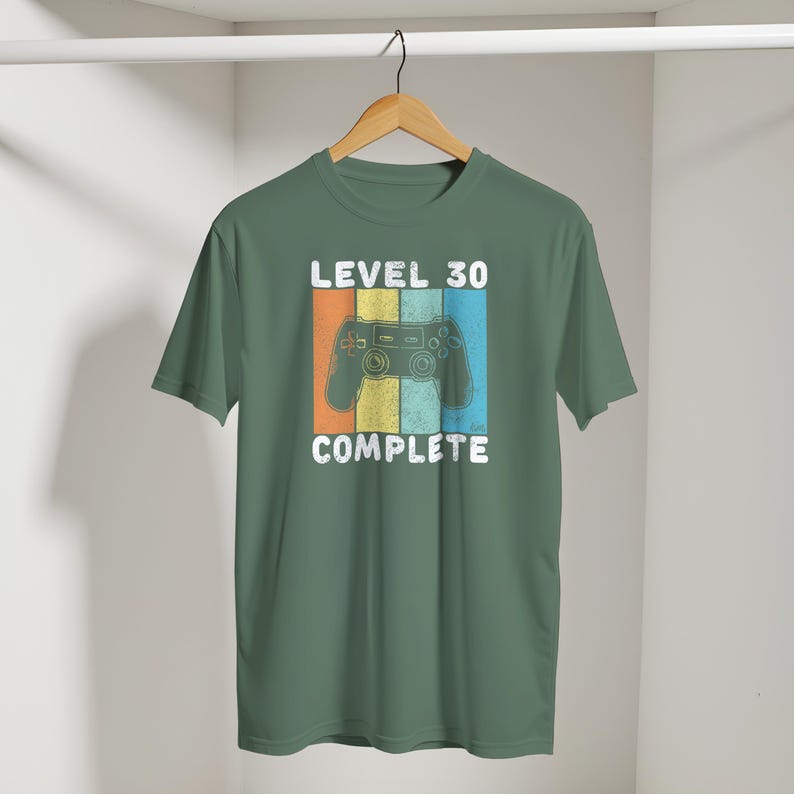 May include: Green t-shirt with a retro design featuring a game controller and the text "LEVEL 30 COMPLETE". The design includes vertical stripes in orange, yellow, and blue. The shirt is hanging on a wooden hanger.