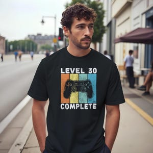 May include: Black t-shirt with the text "LEVEL 30 COMPLETE" and a retro video game controller graphic. The design features orange, yellow, and blue vertical stripes. A casual tee for gamers celebrating a milestone.