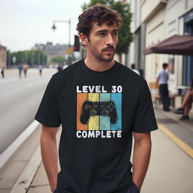 May include: Black t-shirt with the text "LEVEL 30 COMPLETE" and a retro video game controller graphic. The design features orange, yellow, and blue vertical stripes. A casual tee for gamers celebrating a milestone.