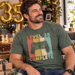 Könnte beinhalten: Ein olivgrünes Kurzarm-T-Shirt mit einem Grafikdruck eines Videospiel-Controllers, zentriert über vier vertikalen Streifen in Rot, Gelb, Beige und Türkis. Der Text lautet LEVEL 35 COMPLETE in einer fetten, abgenutzten Retro-Schriftart.