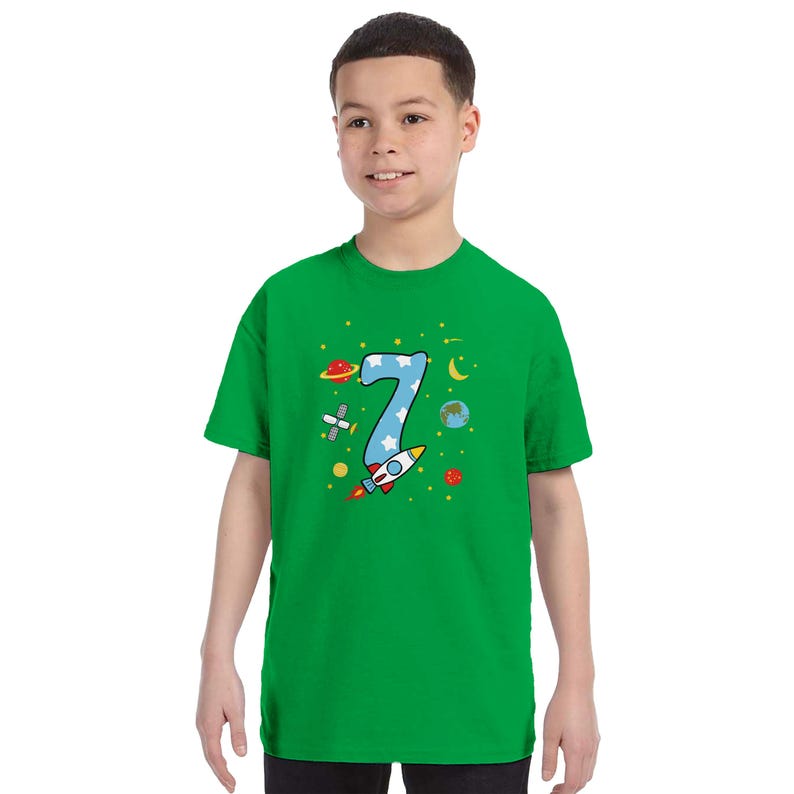 May include: A green t-shirt featuring a number 7 in blue with white stars, a rocket ship, planets, and other space-themed graphics. The shirt is a short-sleeved crew neck.