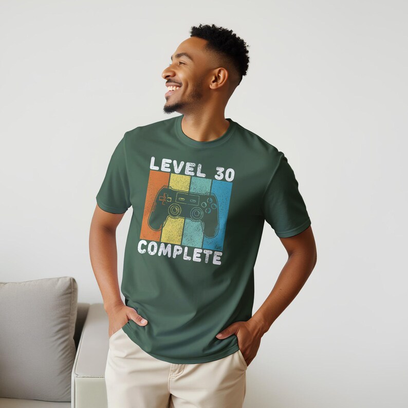 May include: Dark green t-shirt with a retro design featuring a video game controller and the text "LEVEL 30 COMPLETE" in white. The controller is set against a vertical striped background of orange, yellow, and blue.
