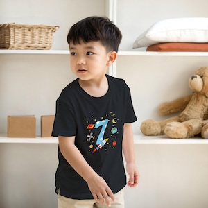May include: Black t-shirt featuring a colorful space-themed design with the number 7. The graphic includes a blue number 7 with stars, planets, and a rocket ship. The shirt is made of a soft material.
