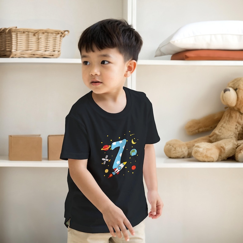 May include: Black t-shirt featuring a colorful space-themed design with the number 7. The graphic includes a blue number 7 with stars, planets, and a rocket ship. The shirt is made of a soft material.