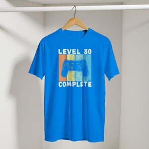 May include: A bright blue t-shirt with the words "LEVEL 30 COMPLETE" in white. A retro-style video game controller graphic is centered on the shirt, with orange, yellow, and blue stripes behind it. The shirt is hanging on a wooden hanger.