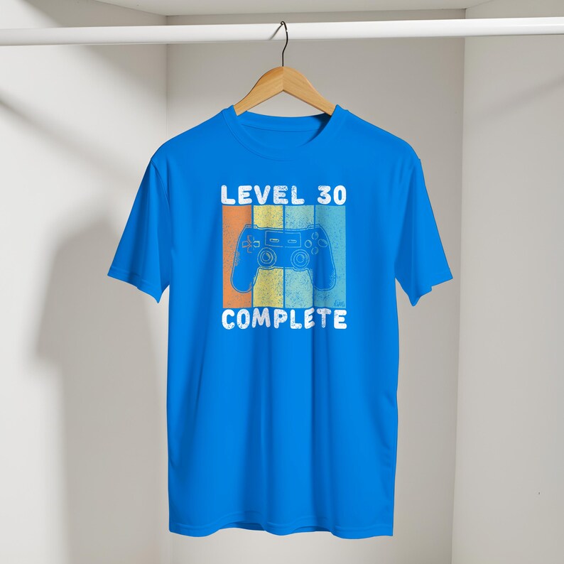 May include: A bright blue t-shirt with the words "LEVEL 30 COMPLETE" in white. A retro-style video game controller graphic is centered on the shirt, with orange, yellow, and blue stripes behind it. The shirt is hanging on a wooden hanger.