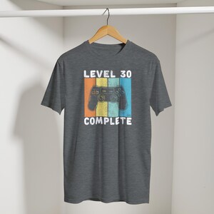 May include: A gray t-shirt with a retro design featuring a video game controller and the text "LEVEL 30 COMPLETE." The design includes orange, yellow, and blue stripes. The shirt is hanging on a wooden hanger.