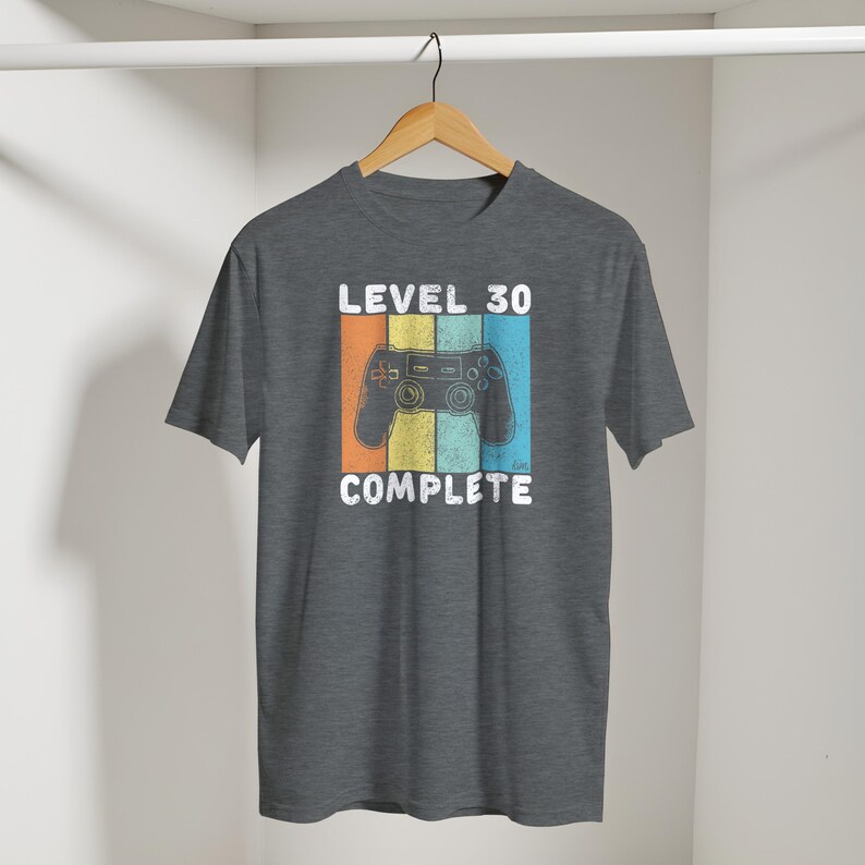 May include: A gray t-shirt with a retro design featuring a video game controller and the text "LEVEL 30 COMPLETE." The design includes orange, yellow, and blue stripes. The shirt is hanging on a wooden hanger.