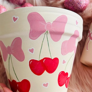 May include: A cream-colored ceramic planter with pink bows and red cherries painted on it. The planter is decorated with small pink hearts.