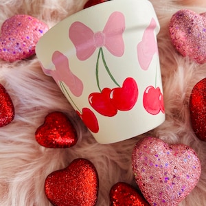 May include: A white ceramic planter with a hand-painted design of pink bows and red cherries. The planter is surrounded by red and pink glitter hearts.