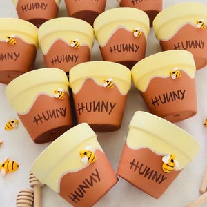 May include: A collection of terracotta-colored flower pots with a yellow honey-like design at the top. The word "HUNNY" is written on each pot. Each pot has a small, decorative bee attached. Wooden honey dippers are also visible.