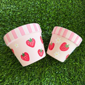May include: Two light pink terracotta plant pots of different sizes, each featuring a hand-painted pattern of red strawberries and small white flowers, with a rim decorated in alternating vertical stripes of light pink and white.