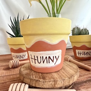 May include: Three hand-painted terracotta flower pots with a yellow and orange design, each labeled "HUNNY" in white. The pots are on a wooden surface, with wooden honey dippers. One pot contains a succulent, another a cactus, and the third, yellow flowers.