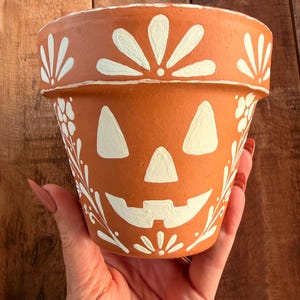 May include: A terracotta clay flower pot painted with white designs. The pot features a jack-o'-lantern face in the center, surrounded by white floral and leaf patterns. The rim is decorated with a repeating white petal motif.