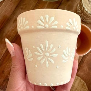 May include: A small, peach-colored terracotta flower pot with white floral designs. The pot features a repeating pattern of stylized flowers and dots around the rim and on the body. The pot is designed for planting flowers or herbs.