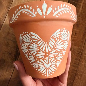 May include: Terracotta-colored flower pot with a white heart design and decorative border. The heart is composed of stylized floral elements. The pot is held by a person. The background is a wooden surface.