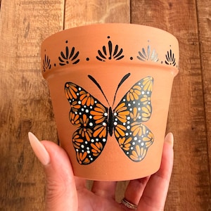 May include: A terracotta plant pot featuring a hand-painted orange and black butterfly design on the front. The butterfly wings are decorated with small white dots. A row of black painted fan-shaped motifs encircles the upper rim of the pot.