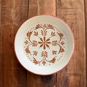 May include: A round, off-white ceramic plate with a brown floral design in the center. The rim is a warm brown color. The plate is set against a wooden background.