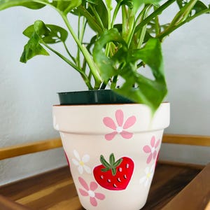 May include: A potted plant with green leaves sits in a beige flower pot. The pot is decorated with a red strawberry and pink and white flower designs. The pot is sitting on a wooden tray.