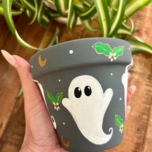 May include: A gray painted ceramic flower pot with a white ghost design. The pot is decorated with gold moons, white dots, and green and white floral accents. The pot is held in a hand.