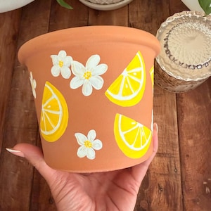 May include: A terracotta-colored plant pot decorated with hand-painted yellow lemon slices and white flowers. The pot is held in a hand, showcasing the design. The pot is designed for indoor or outdoor use.