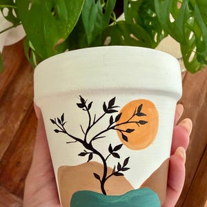 May include: A white ceramic plant pot with a hand-painted design. The design features a black tree silhouette, an orange sun, and abstract shapes in brown and teal. The pot is designed for indoor plants.