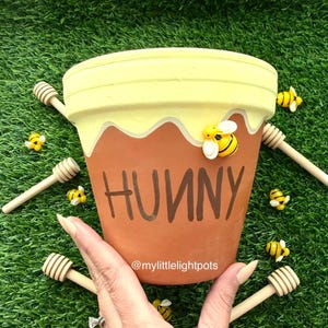 May include: A terracotta flower pot painted with a yellow honey design and the word "HUNNY". The pot is decorated with small bee figurines and honey dippers. The pot is held against a green background.