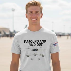Puede incluir: Camiseta gris claro con el texto "F AROUND AND FIND OUT" en negro, junto con una ilustración de varios aviones de combate. La camiseta también presenta un pequeño logotipo "Red, White, and Wit" con una bandera estadounidense.
