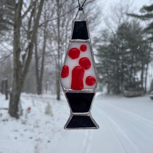 May include: A stained glass lava lamp ornament. The ornament features a black top and bottom, a white center, and red lava-like shapes. The ornament is hanging against a snowy background.