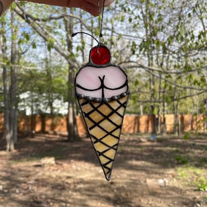 May include: A stained glass ice cream cone ornament with a pink ice cream scoop and a red cherry on top. The cone is yellow with black grid lines.