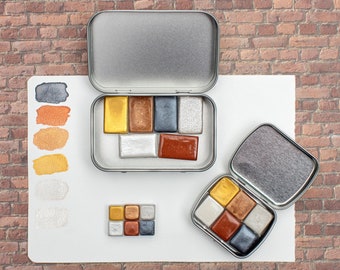 May include: Two metal watercolor paint palettes with a variety of metallic colors, including gold, silver, copper, and bronze. The palettes are open and show the individual paint pans.