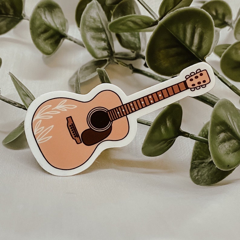 Pode incluir: Um adesivo de guitarra ac&uacute;stica cor p&ecirc;ssego com um design floral branco. A guitarra tem um bra&ccedil;o marrom e um furo de som marrom.
