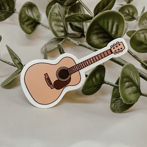 Pode incluir: Um adesivo de uma guitarra ac&uacute;stica marrom claro com um furo de som marrom escuro. A guitarra tem um contorno branco e est&aacute; sobre um fundo branco.