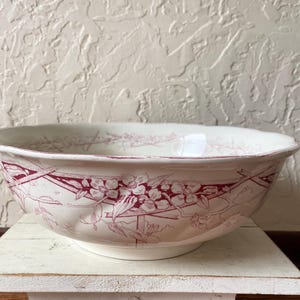 May include: A round ceramic bowl with a cream-colored base and a deep red floral pattern featuring blossoms and branches wrapping around the exterior. The bowl has a scalloped rim and a smooth, glossy finish, showcasing a detailed botanical design.