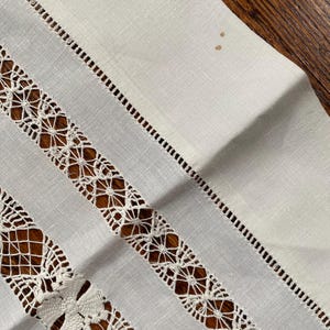 May include: Close-up of a white linen table runner with intricate, openwork lace and decorative stitching. The runner features alternating stripes of solid fabric and detailed lace patterns, creating a textured and elegant design. The background is a dark wood surface.