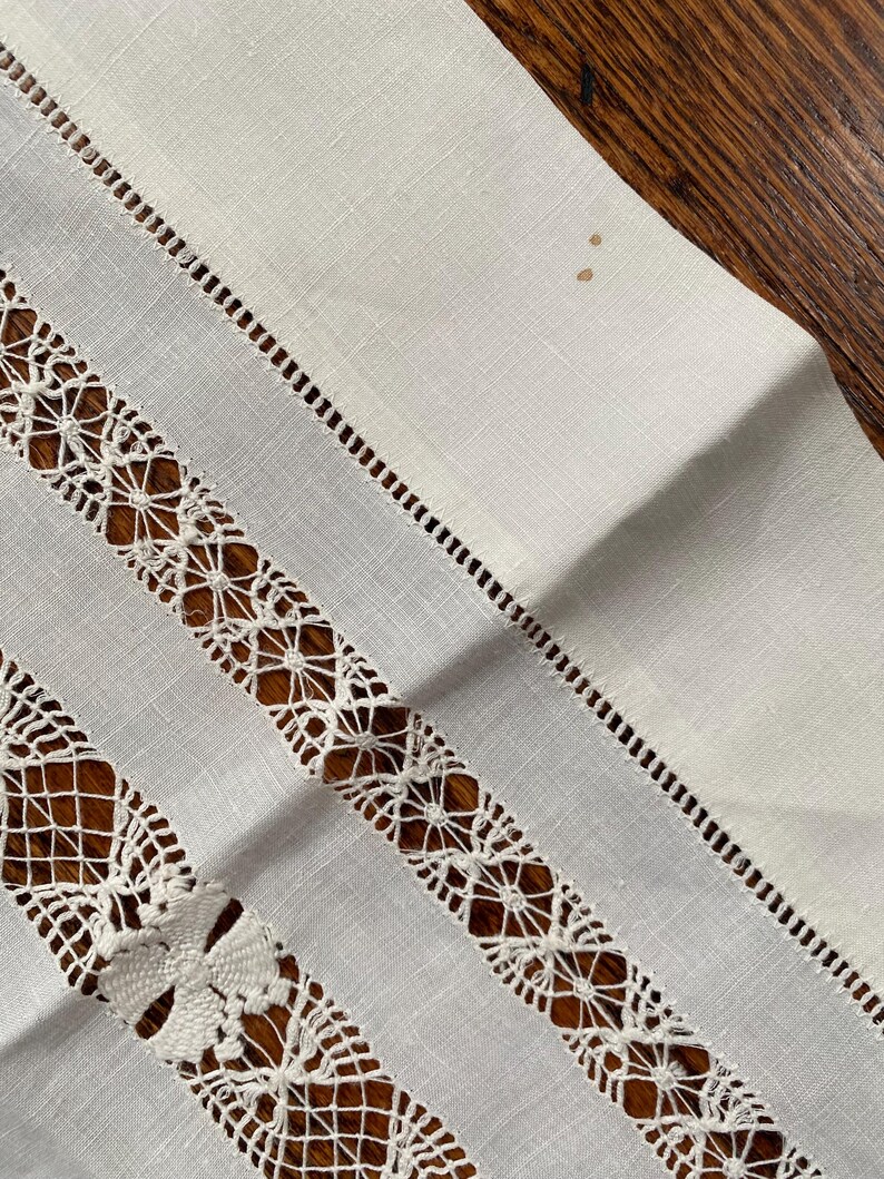 May include: Close-up of a white linen table runner with intricate, openwork lace and decorative stitching. The runner features alternating stripes of solid fabric and detailed lace patterns, creating a textured and elegant design. The background is a dark wood surface.