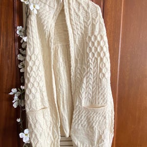 May include: An off-white open-front cardigan featuring a complex cable knit pattern throughout. The garment includes two front patch pockets and a fringed hemline. The knit texture consists of various vertical panels of braided and honeycomb designs.