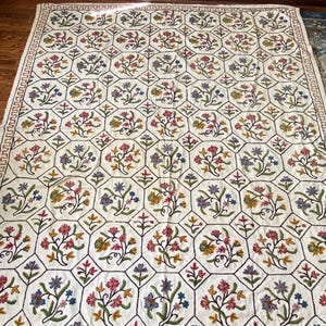 May include: A rectangular off-white textile featuring a repeating hexagonal grid pattern outlined in dark thread. Each hexagon contains a colorful embroidered floral motif in shades of red, blue, yellow, and green, with a decorative border.