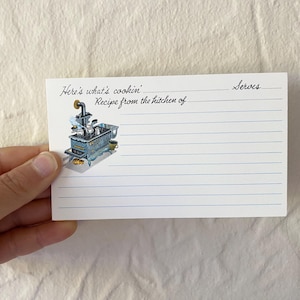 May include: A white rectangular recipe card with blue horizontal lines. The text reads Here's what's cookin', Recipe from the kitchen of, and Serves. A small illustration of a blue vintage wood-burning stove with pots sits on the left side.