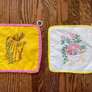 May include: Two square fabric potholders with embroidered designs. The left potholder is yellow with a pink border and features a brown teapot with green vines. The right potholder is white with a yellow border and a teacup with pink flowers.