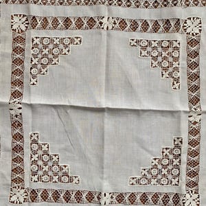 May include: White linen tablecloth with intricate lace detailing. The tablecloth features a central panel with corner floral designs and a border of geometric and floral lace patterns. A vintage textile.
