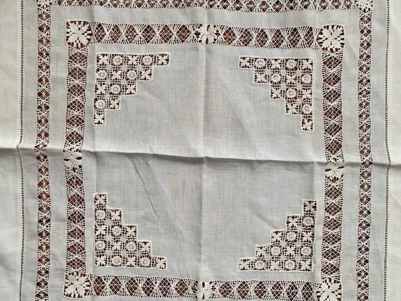 May include: White linen tablecloth with intricate lace detailing. The tablecloth features a central panel with corner floral designs and a border of geometric and floral lace patterns. A vintage textile.