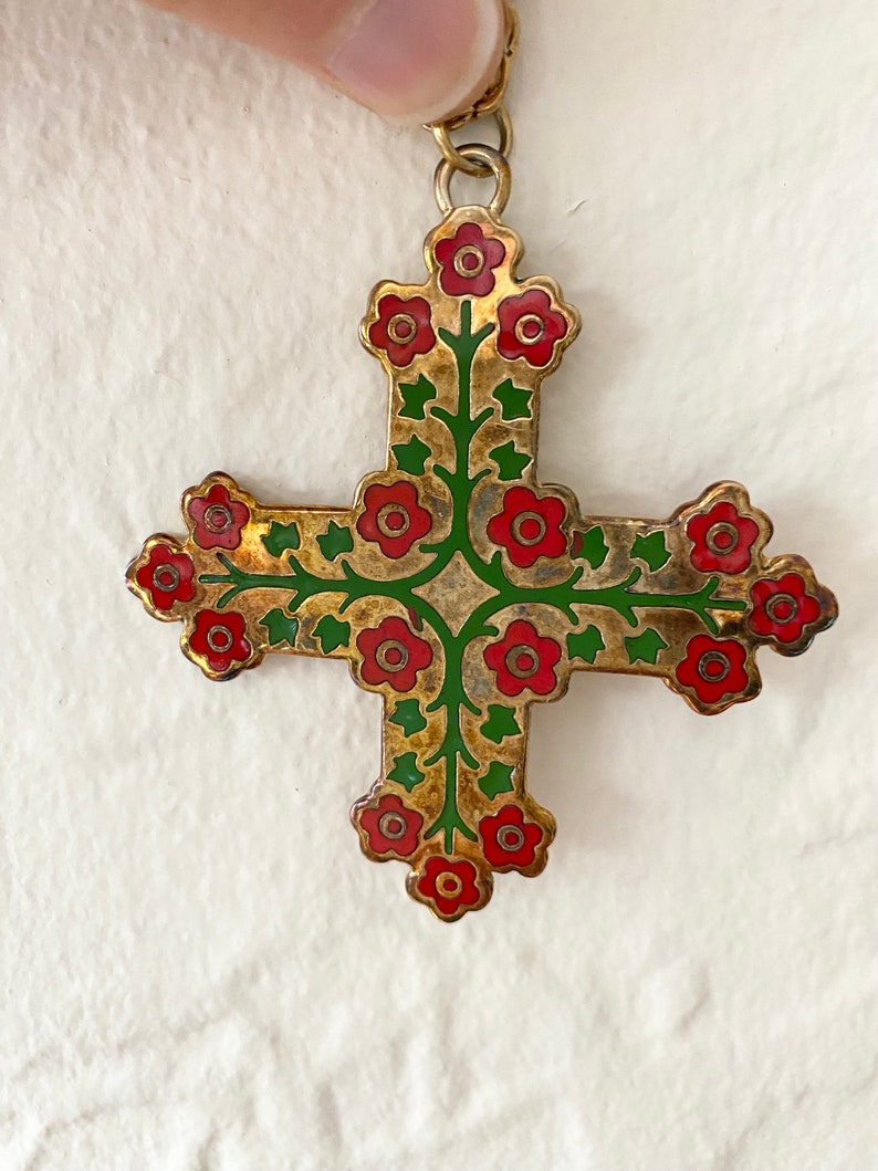 May include: A gold-toned cross pendant with red and green enamel detailing. The cross is decorated with a floral design featuring red flowers and green leaves.