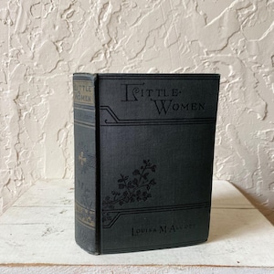 May include: A dark green hardcover book titled Little Women by Louisa M. Alcott. The front cover features black embossed lettering and a decorative floral branch design. The spine displays the title and author name in black text with gold accents.