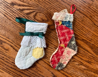 May include: Two small, handmade Christmas stockings. One is light blue with a yellow patch and a green bow. The other is red with a patchwork design and a red ribbon loop. Both have lace trim.