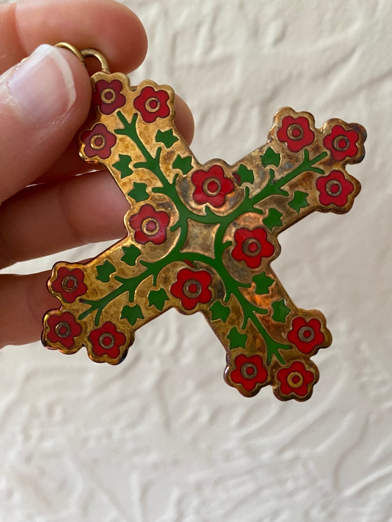 May include: A gold-toned metal cross pendant with a floral design. The cross is decorated with red and green enamel, featuring stylized flowers and leaves.